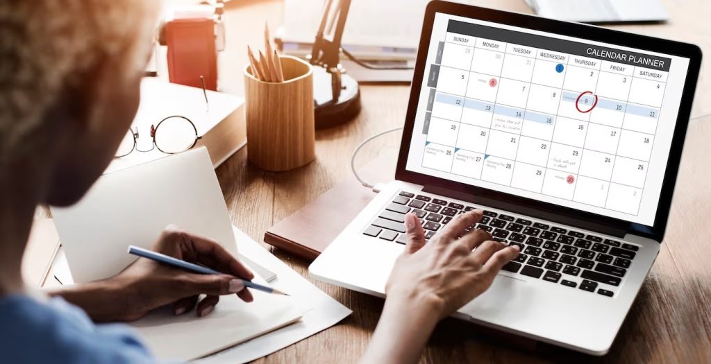 A person working at a desk, typing on a laptop displaying a digital calendar planner while writing in a notebook